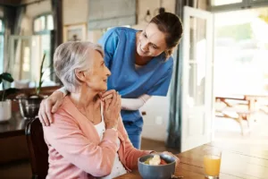 Une femme âgée est assise à table, sourit et regarde une aide-soignante vêtue de bleu qui lui pose une main réconfortante sur l'épaule. Elle est heureuse d'avoir pu bénéficier du service de repas à domicile de Liège.