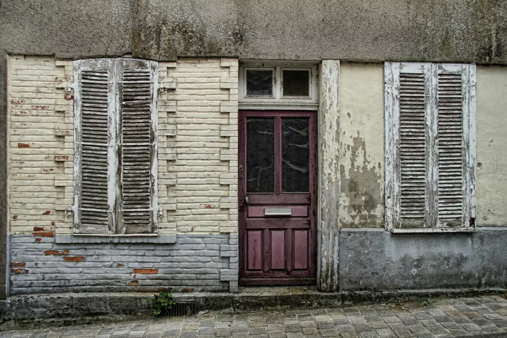 Façade d'une maison ancienne et délabrée avec volets et porte en mauvais état. Cette image illustre les conditions pouvant donner droit à l'Allocation de relogement en Belgique.