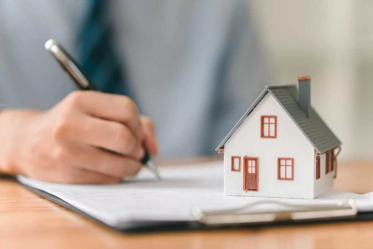 Signature d’un document lié au logement avec une maquette de maison, illustrant le rôle des Associations de Promotion du Logement (APL) dans l’accompagnement des citoyens pour l’accès à un habitat décent.