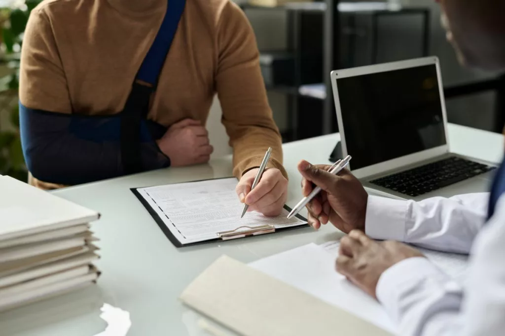 Un patient avec un bras en écharpe remplit des documents avec l'aide d'un professionnel, illustrant une demande d’indemnisation auprès du Fonds des Accidents Médicaux en Belgique.