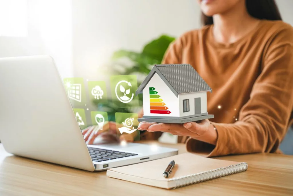 Une femme tenant une maison miniature avec un label énergétique, illustrant la prime communale Audit Logement de la Ville de Namur, une aide financière pour améliorer la performance énergétique des habitations.