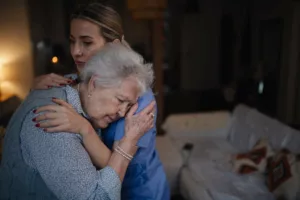 Une femme âgée en détresse est réconfortée par une proche dans un salon sombre, illustrant la douleur liée au décès d'une personne handicapée.