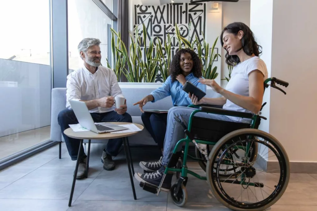 Discussion entre collègues sur les démarches pour obtenir la Prime rattrapage, avec une femme en fauteuil roulant dans un environnement de travail inclusif en Belgique.